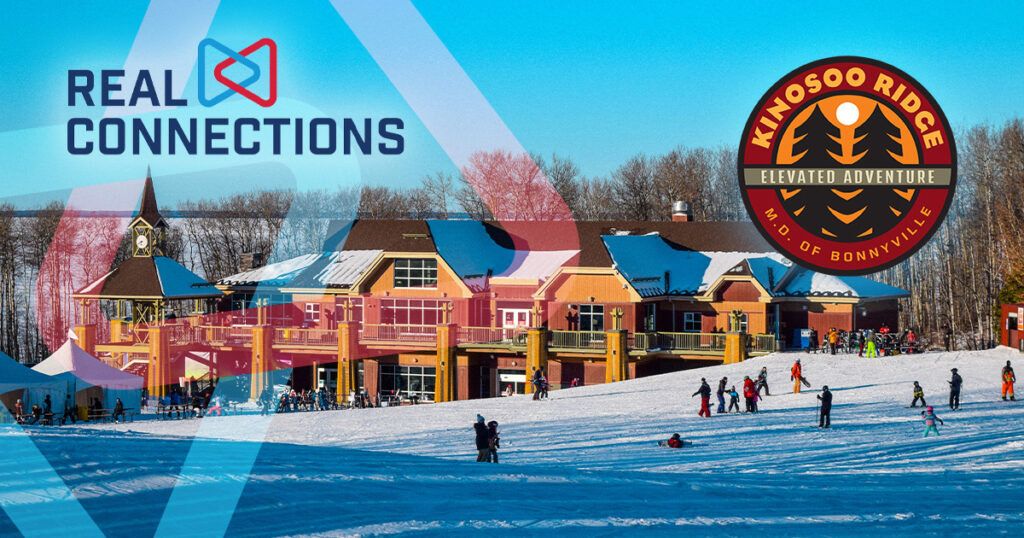 Real Connections logo and Kinosoo Ridge Resort M.D. of Bonnyville Resort logo. Kinosoo Ridge Resort Chalet with skiers on a snowy hill in Cold Lake, Alberta. Blue sky and a frozen lake behind.