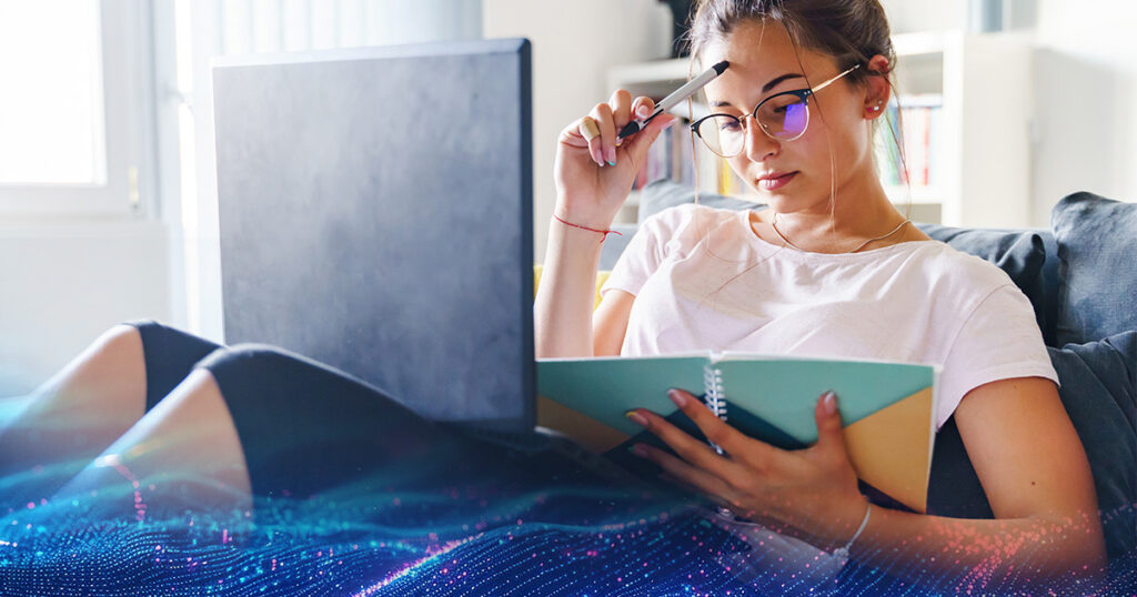 A young woman sitting on the couch at home and studying with a notebook and a laptop on her lap.