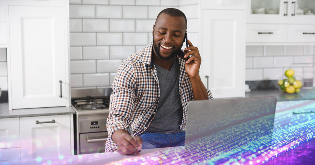 A man on the phone in the kitchen in front of a laptop, taking notes.