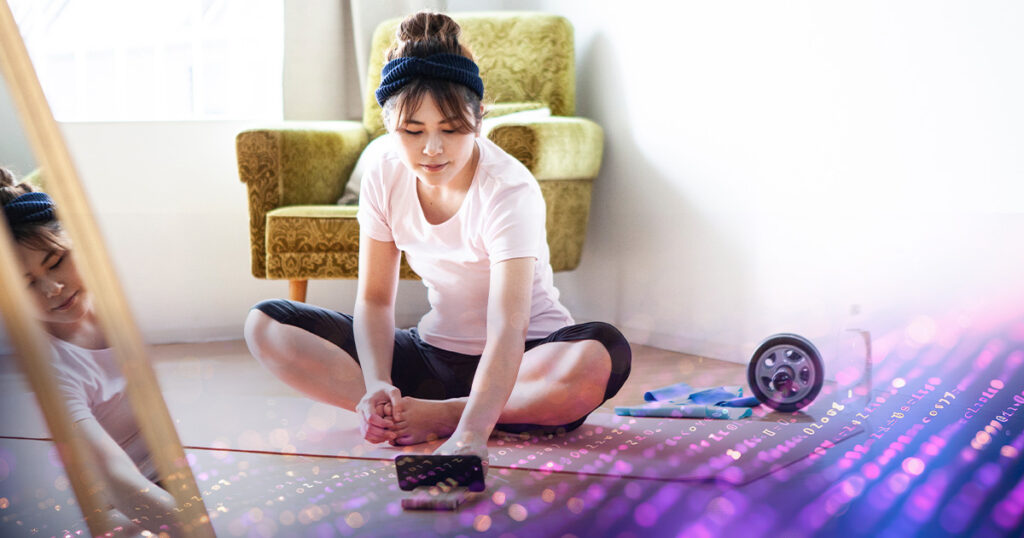 Young woman sitting on a yoga mat on the floor at home and using her cell phone.