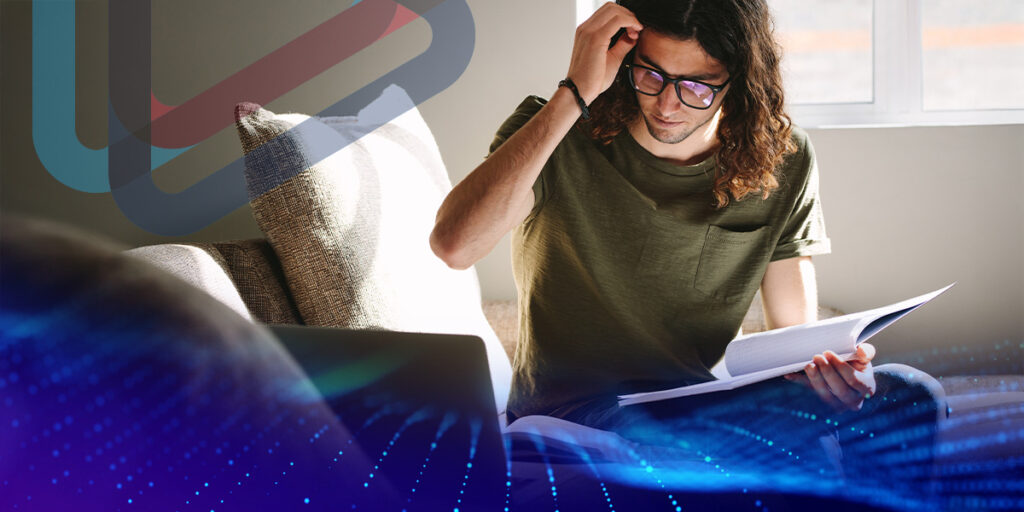 Image of a male student, wearing glasses, holding a book and sitting on a coush with a laptop beside him.