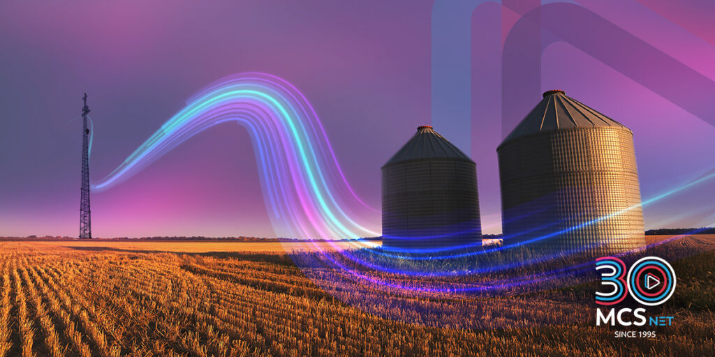 Image of a two grain bins in a field with an internet tower nearby and a digital image of a flowing wave extending from the tower. MCSnet's icon is also in the background.