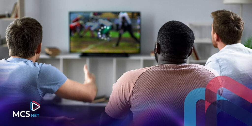 The view from behind a seating area of three young men watching a sports event on TV and the TV signal is buffering.
