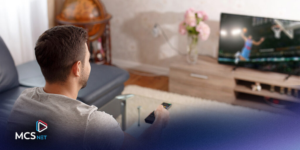 View from behind a young man sitting on a couch, watching the TV in front of him and holding a remote.