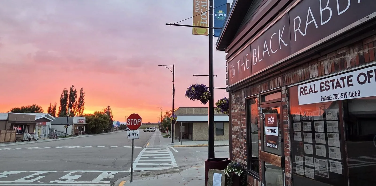 Main street in Boyle, Alberta at sunset, with a Boyle banner on a lamp post, hanging flower baskets, a four way stop and storefronts running down the block