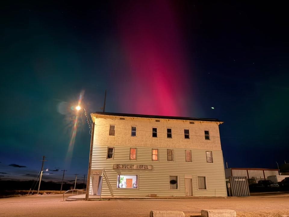 The three storey Derwent Hotel building in Derwent, Alberta, lit by a street light at night under green and red northern lights