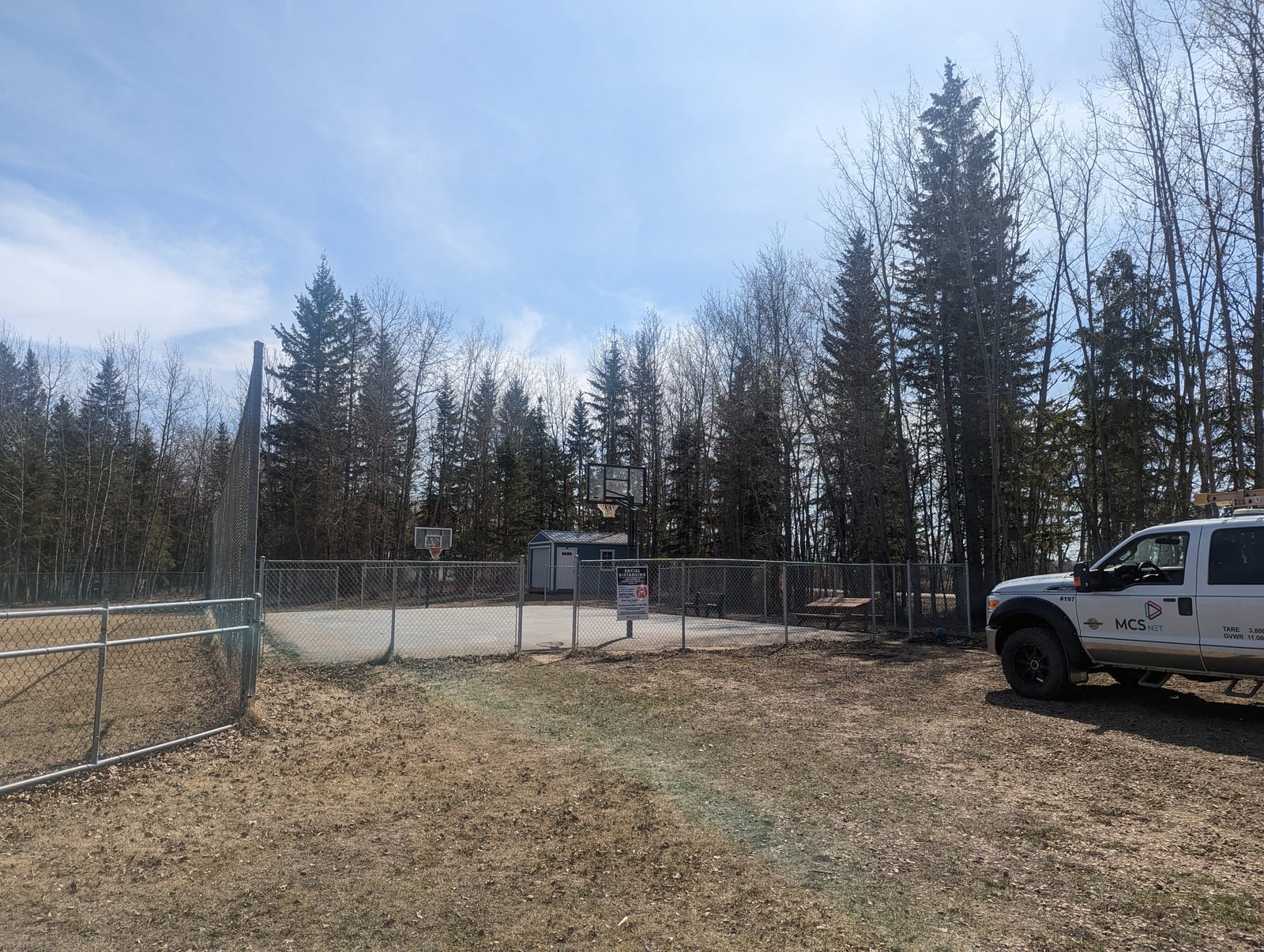 A fenced sport court with basketball hoops, a small shed, and picnic tables in the trees at Island Lake Memorial Park, with an MCSnet service truck parked alongside