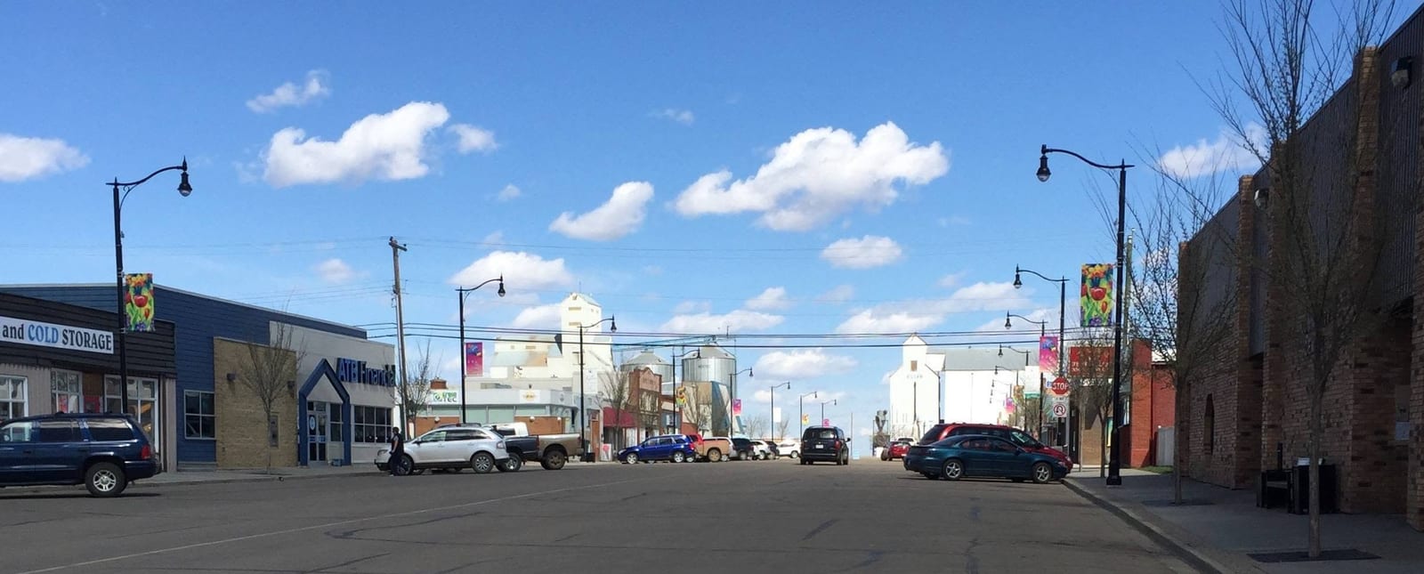Main street in Killam, Alberta on a clear spring day, with brick storefronts, banner flags on the lamp posts, angle parked vehicles and grain elevators at the far end of the street