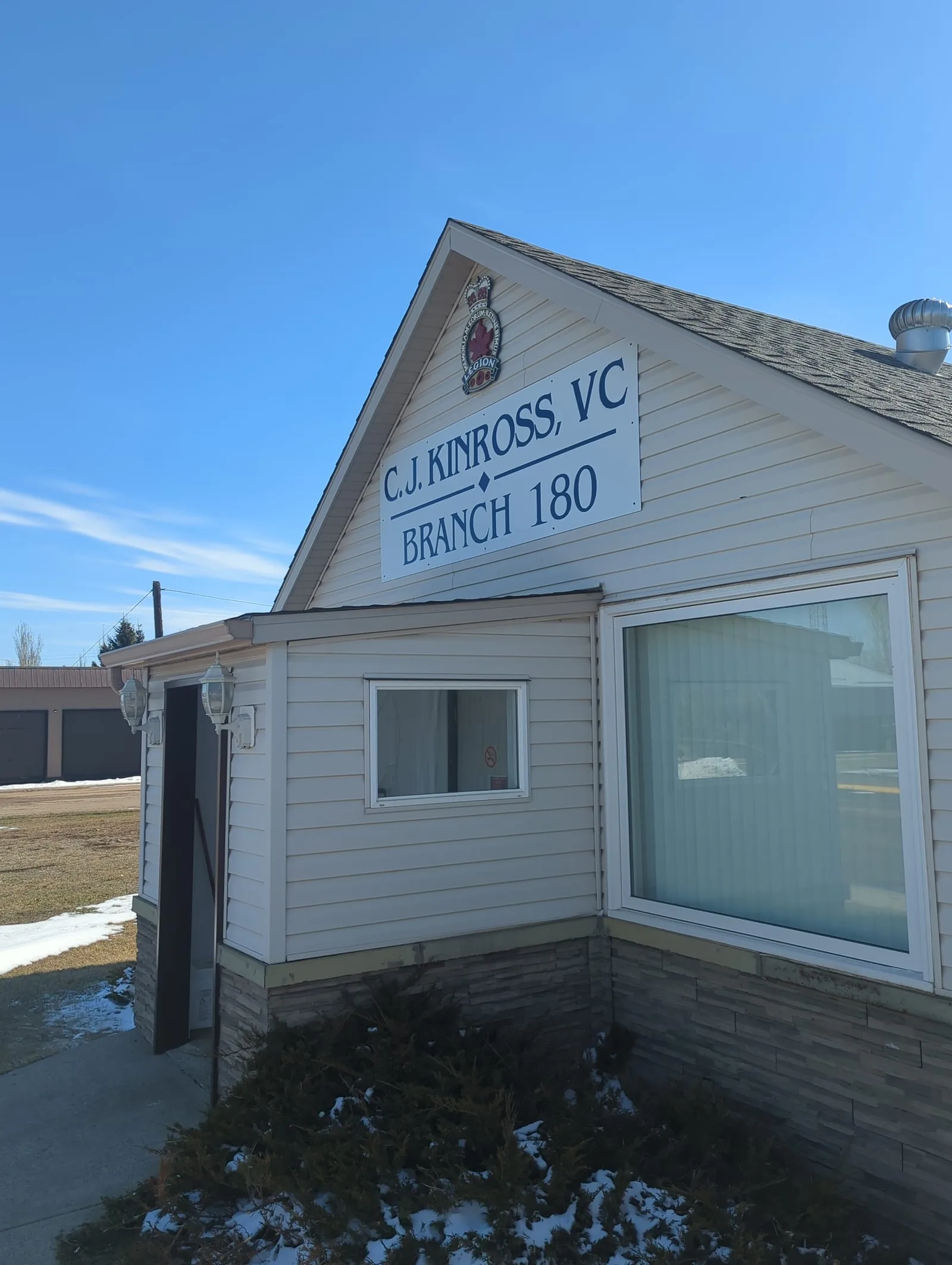 Front gable of the C.J. Kinross VC Royal Canadian Legion Branch 180 hall in Lougheed, Alberta, with the branch sign and crest under a clear blue sky
